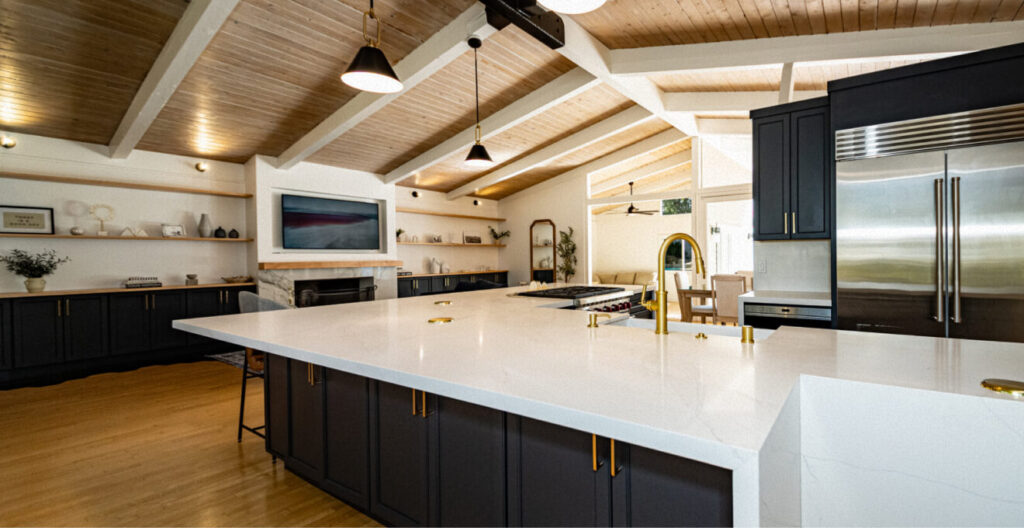 Spacious modern kitchen with vaulted wooden ceiling, large marble island, black cabinets, stainless steel appliances, and warm lighting.