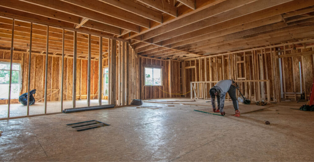 Spacious construction site with wooden frames and beams. A worker measures the floor, indicating progress in a calm, industrious atmosphere.
