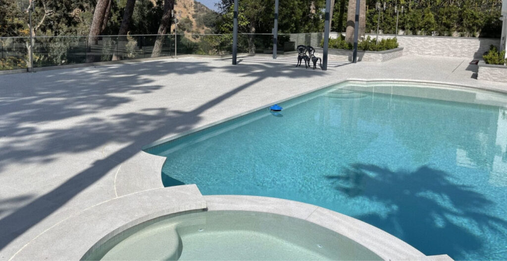 A serene outdoor pool area with a clear blue pool and adjacent jacuzzi. Sunlight casts tree shadows on the white deck. Two black chairs sit in the background.